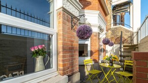 A small outdoor seating area with yellow chairs and a table, a window with a vase of pink flowers, and a brick wall with a hanging purple flower. - 2 Bed in Broadstairs (oc-th017) (Broadstairs)