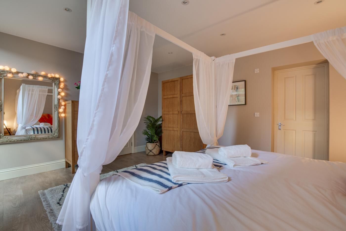 A bedroom with a four-poster bed, a large mirror with lights, a wooden wardrobe, and a door.