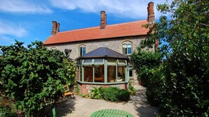 Une maison en pierre avec un toit recouvert de tuiles rouges, une véranda avec des portes vitrées et un jardin agrémenté d’un banc et d’une table en bois.