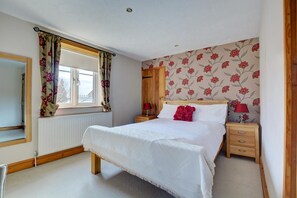 A bedroom with a large bed, a wooden nightstand, a mirror, and a window with floral curtains. - 2 Bed in Bodham (oc-1303) (Bodham)