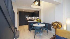 A modern kitchen with dark cabinetry, a central island, and a dining area with a round table and blue chairs. - 2 Bed in Bath (oc-bhr8) (Bath)
