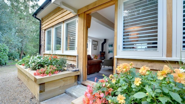 A cozy cottage with a wooden exterior, large windows with shutters, a visible fireplace, and a garden with flowers and a shoe. - 1 Bed in Cranbrook (oc-cb652) (Cranbrook)