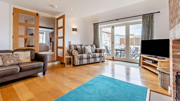 A spacious living room with a wooden floor, a sofa, a striped armchair, and a television on a wooden stand. - 4 Bed in Ringstead (oc-1672) (Ringstead)