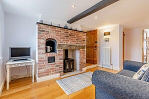 A living room with a brick fireplace, a television on a white table, a sofa, and a wooden floor. - 4 Bed in Ringstead (oc-1672) (Ringstead)