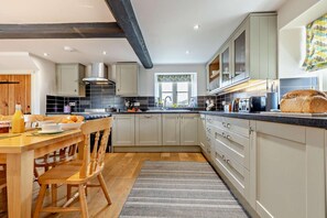 A modern kitchen with a wooden dining table, light-colored cabinets, and a large loaf of bread on the counter. - 4 Bed in Ringstead (oc-1672) (Ringstead)