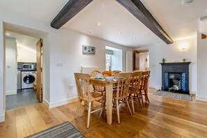 A dining area with a wooden table and chairs, a fireplace, and a kitchen visible in the background. - 4 Bed in Ringstead (oc-1672) (Ringstead)