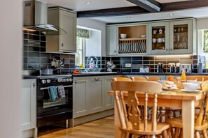 A modern kitchen with grey cabinets, a black stove, and a dining area with wooden chairs. - 4 Bed in Ringstead (oc-1672) (Ringstead)