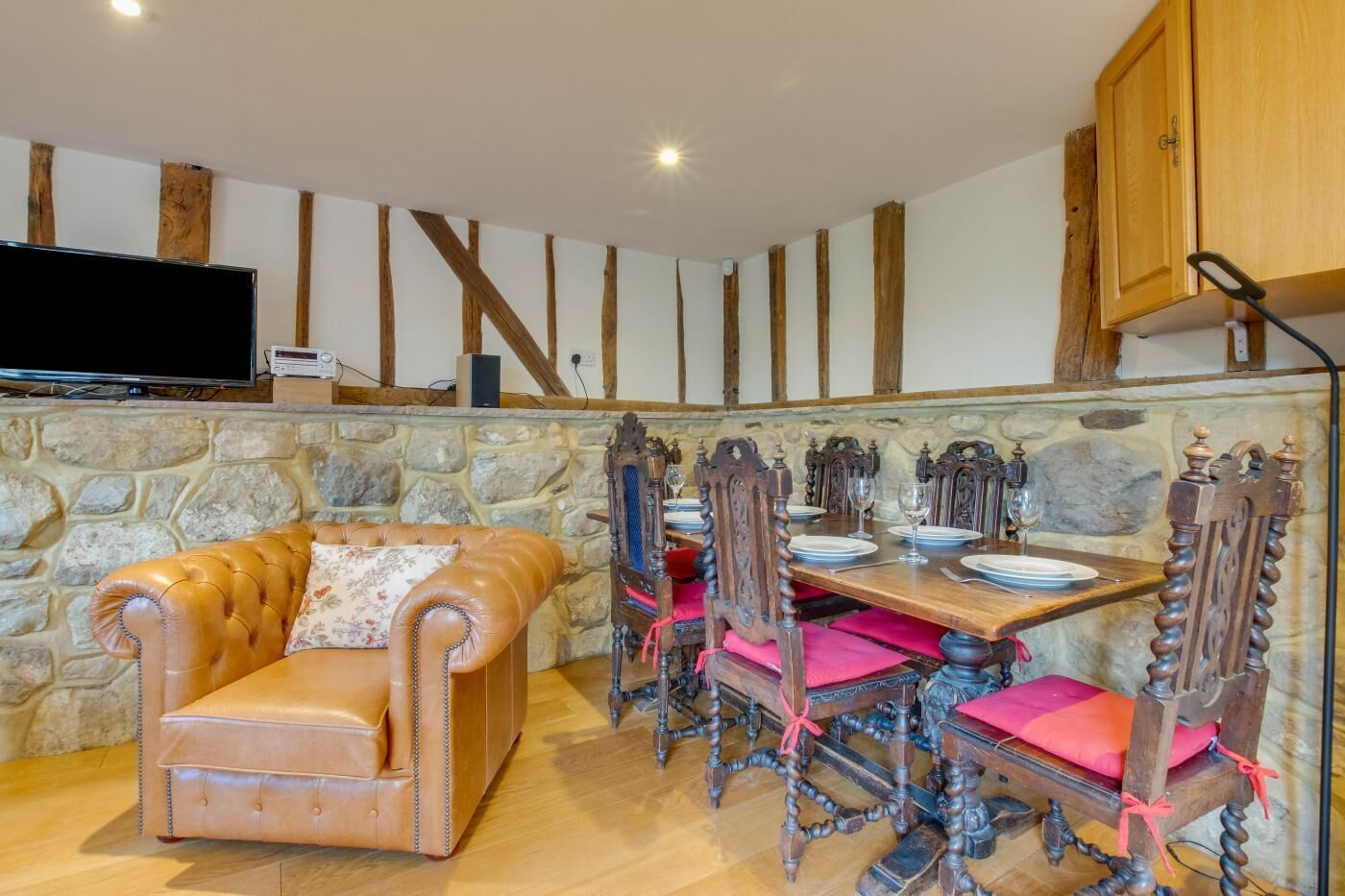 Una habitación acogedora con pared de piedra, vigas de madera, un sofá de cuero, una mesa de comedor de madera con sillas y un televisor en la pared.