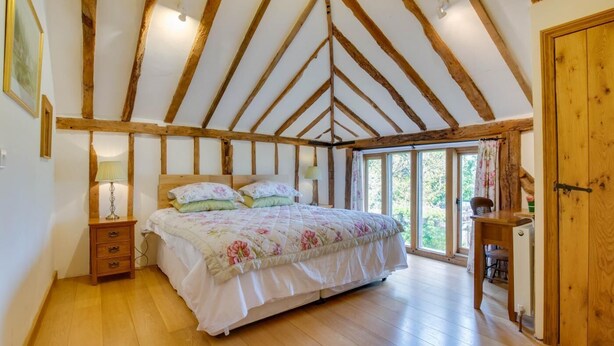 A bedroom with a wooden ceiling, a bed with floral bedding, a bedside table with a lamp, and a view of the outdoors through large windows. - 2 Bed in Cranbrook (oc-cb594) (Cranbrook)