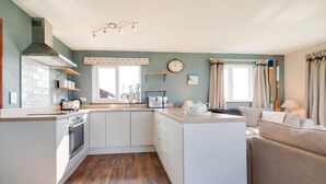 A compact kitchen with white cabinets, a stove, and a sink. There is a dining area with a sofa and a table. - 3 Bed in Eccles Beach (oc-852) (Eccles-on-Sea)