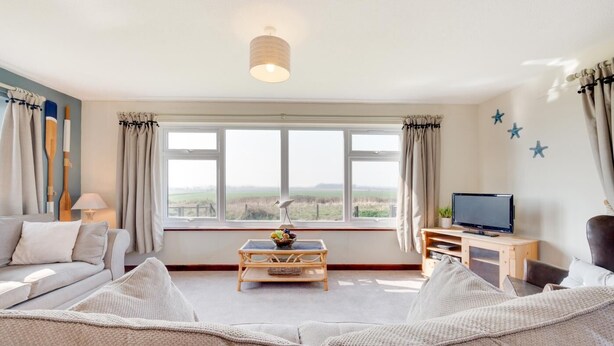 A living room with a sofa, a coffee table, a TV, and a window with a view of the countryside. - 3 Bed in Eccles Beach (oc-852) (Eccles-on-Sea)