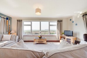 A living room with a sofa, a coffee table, a TV, and a window with a view of the countryside. - 3 Bed in Eccles Beach (oc-852) (Eccles-on-Sea)