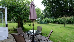 En udendørs terrasse med et rundt bord, fire stole, en parasol og en dør, der fører til haven.