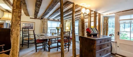 Un interior rústico con vigas de madera a la vista, una zona de comedor con mesa y sillas, y un aparador de madera con cajones.