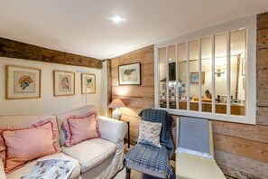 Una sala acogedora con una pared de madera, un sofá blanco, un sillón azul y una zona de comedor visible a través de una ventana.