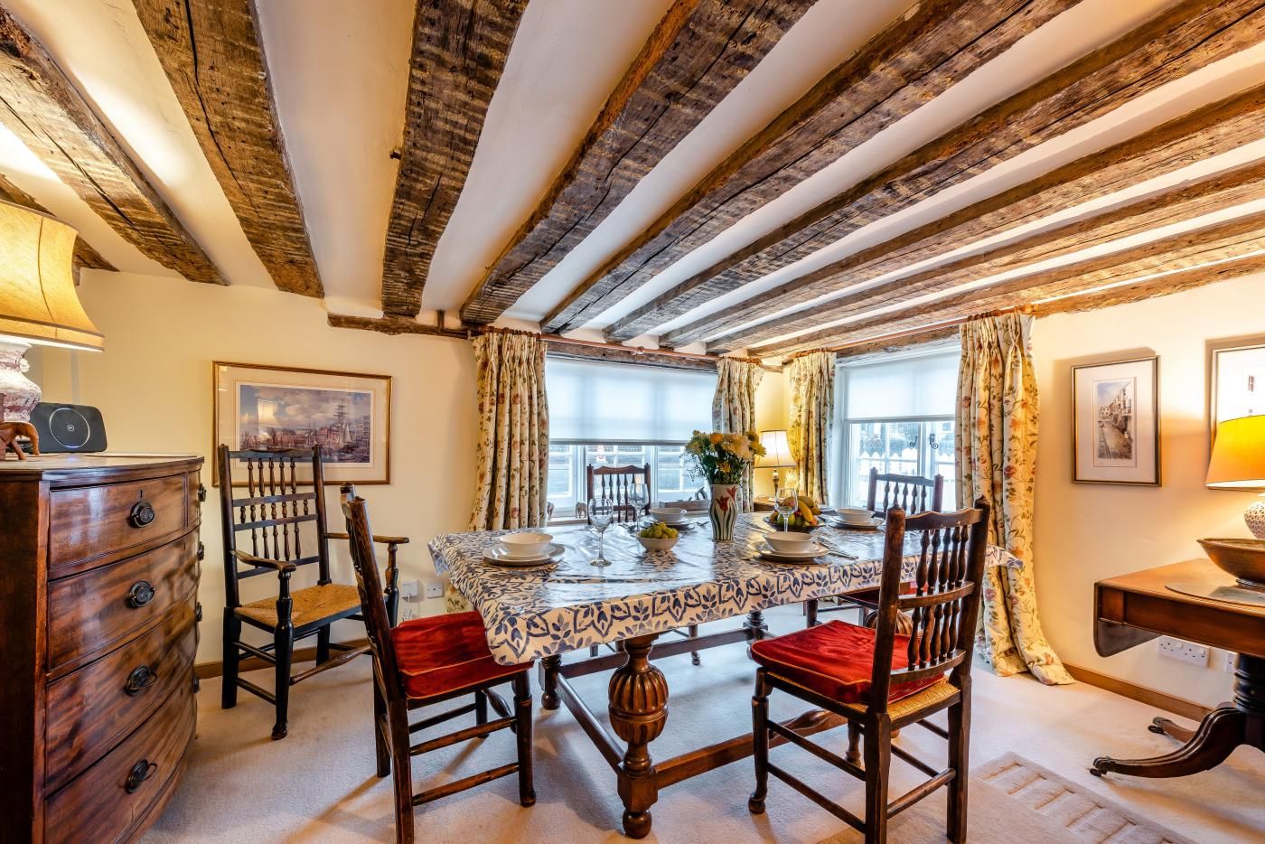 Un coin repas avec une table dressée pour quatre personnes, entouré de chaises en bois avec des coussins rouges, d’une commode en bois et d’une lampe.