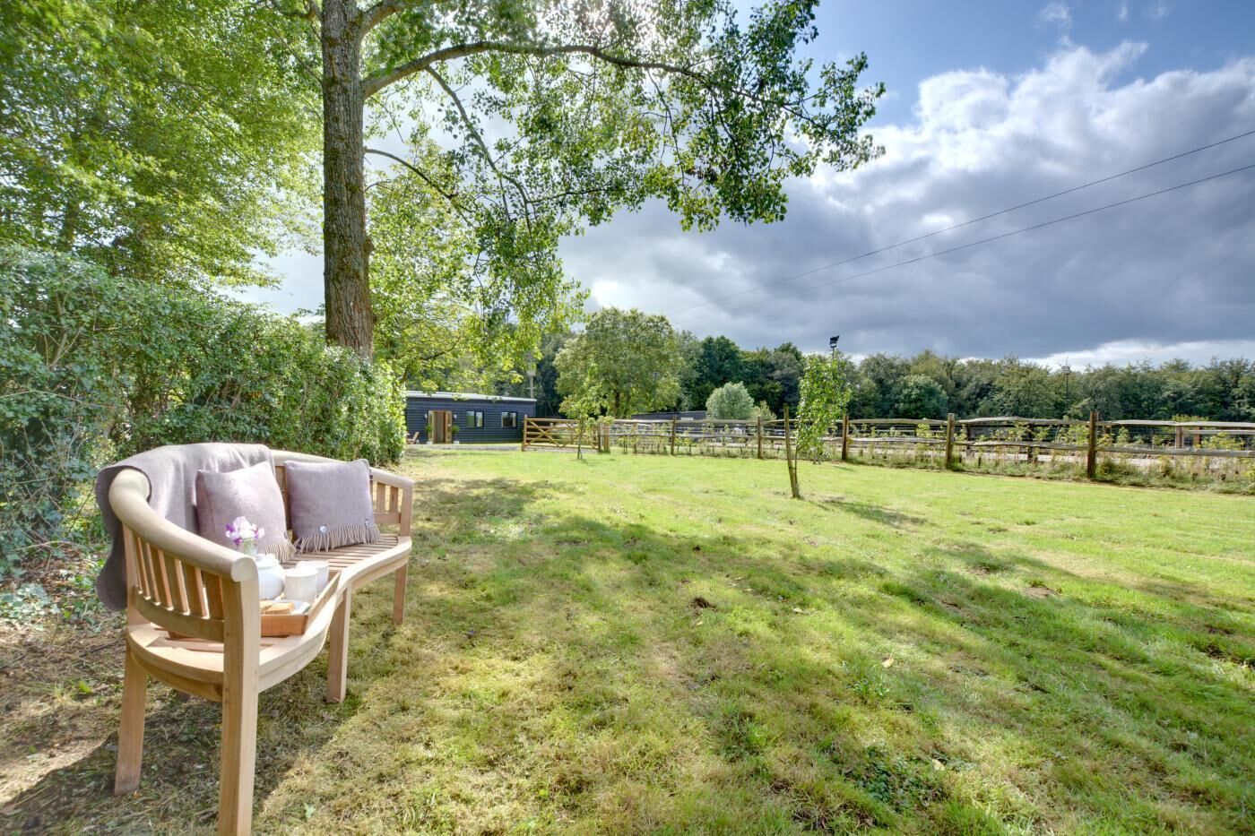 A wooden bench with cushions in a grassy outdoor area with a fence and trees in the background.