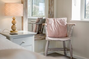Ein Zimmer mit einem weißen Schreibtisch, einer Lampe mit gemustertem Lampenschirm, einem Stuhl mit gestreifter Sitzkissen und einem Fenster mit Vorhängen.