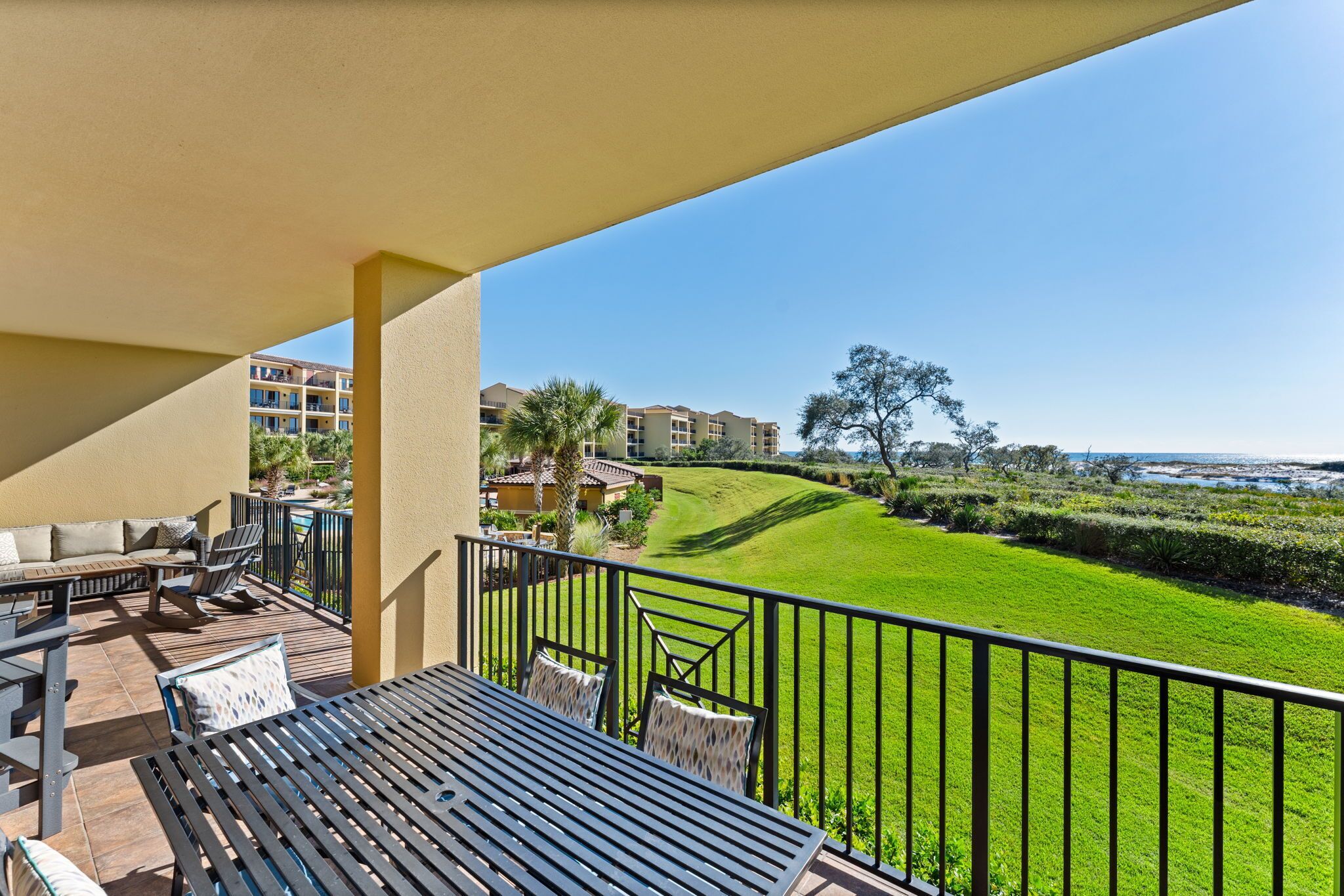 A balcony with outdoor furniture, a view of a well-maintained lawn, and a distant building complex.