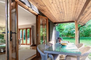 Een overdekt eethoekje buiten met een glazen tafel en rieten stoelen, verbonden met een binnenruimte via een schuifdeur van glas.
