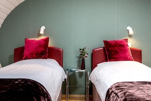 Une chambre d’hôtel avec deux lits, chacun doté d’une tête de lit rouge et de draps blancs. Une petite table avec un vase de fleurs se trouve entre les deux lits.