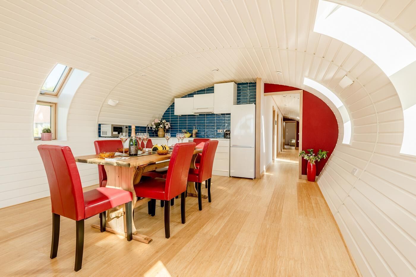 A modern kitchen with a curved ceiling, a dining area with red chairs, and a visible kitchen counter.