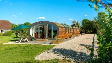 Un bâtiment moderne en bois aux lignes courbes, doté de grandes portes vitrées, d’une allée en gravier et d’une table de pique-nique surmontée d’un parasol vert.