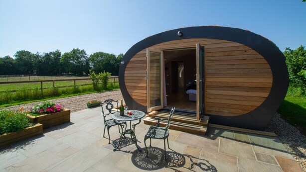 A wooden cabin with a circular design, an open door, a patio with a table and chairs, and a garden area with flowers.