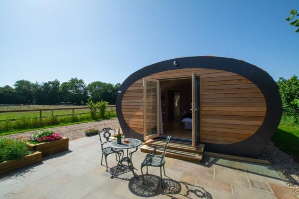 A wooden cabin with a circular design, an open door, a patio with a table and chairs, and a garden area with flowers.