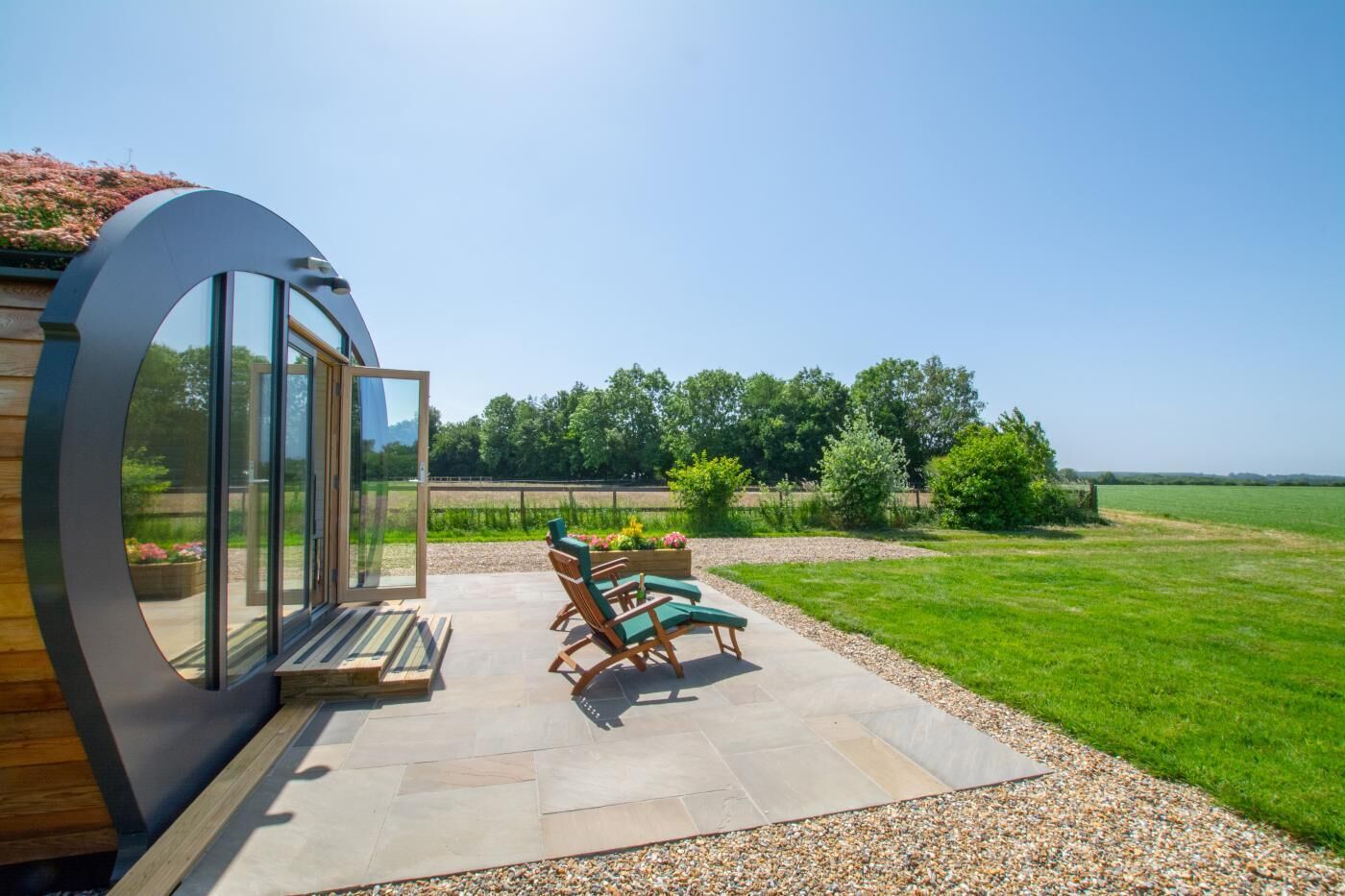A modern outdoor patio with a curved glass wall, a seating area with two lounge chairs, and a well-maintained lawn.