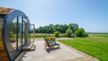 A modern outdoor patio with a curved glass wall, a seating area with two lounge chairs, and a well-maintained lawn.