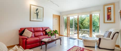 A spacious living room with a red leather sofa, a white armchair, and a coffee table. There is a large window with a view of greenery outside.