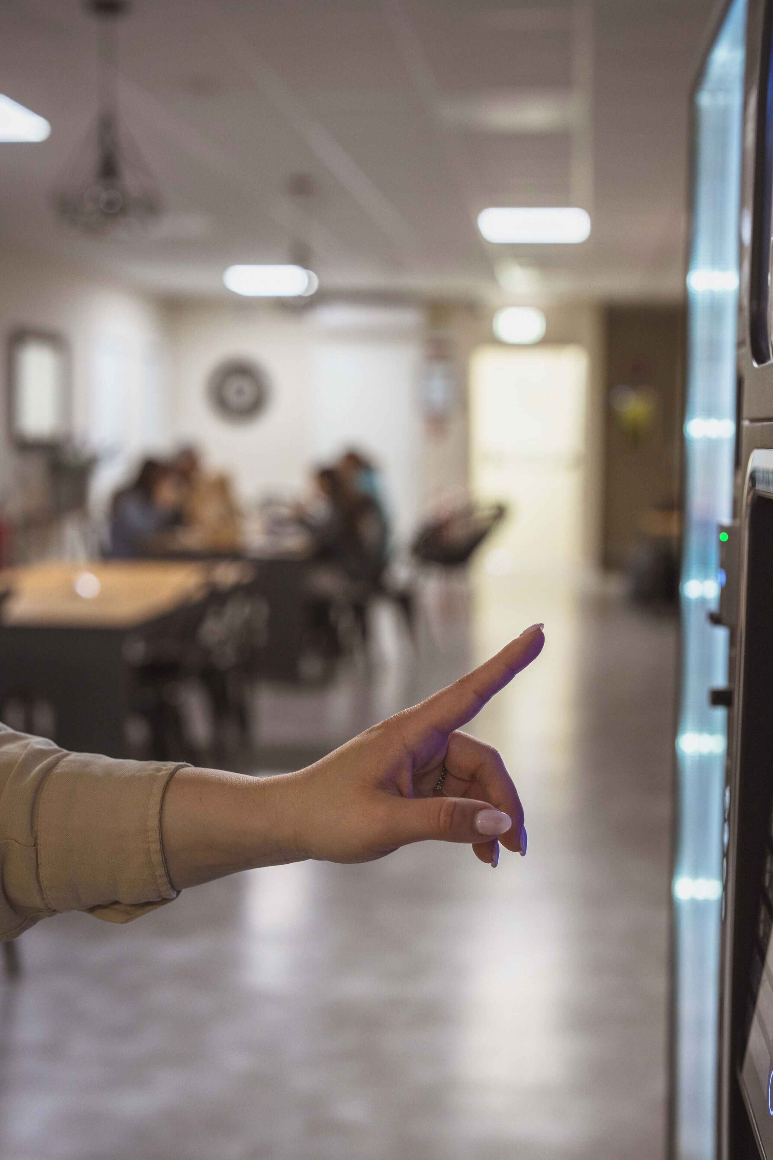 Une main pointant vers un distributeur automatique dans une pièce aménagée avec des tables et des chaises, ce qui suggère un espace de restauration informel ou une salle de réunion.