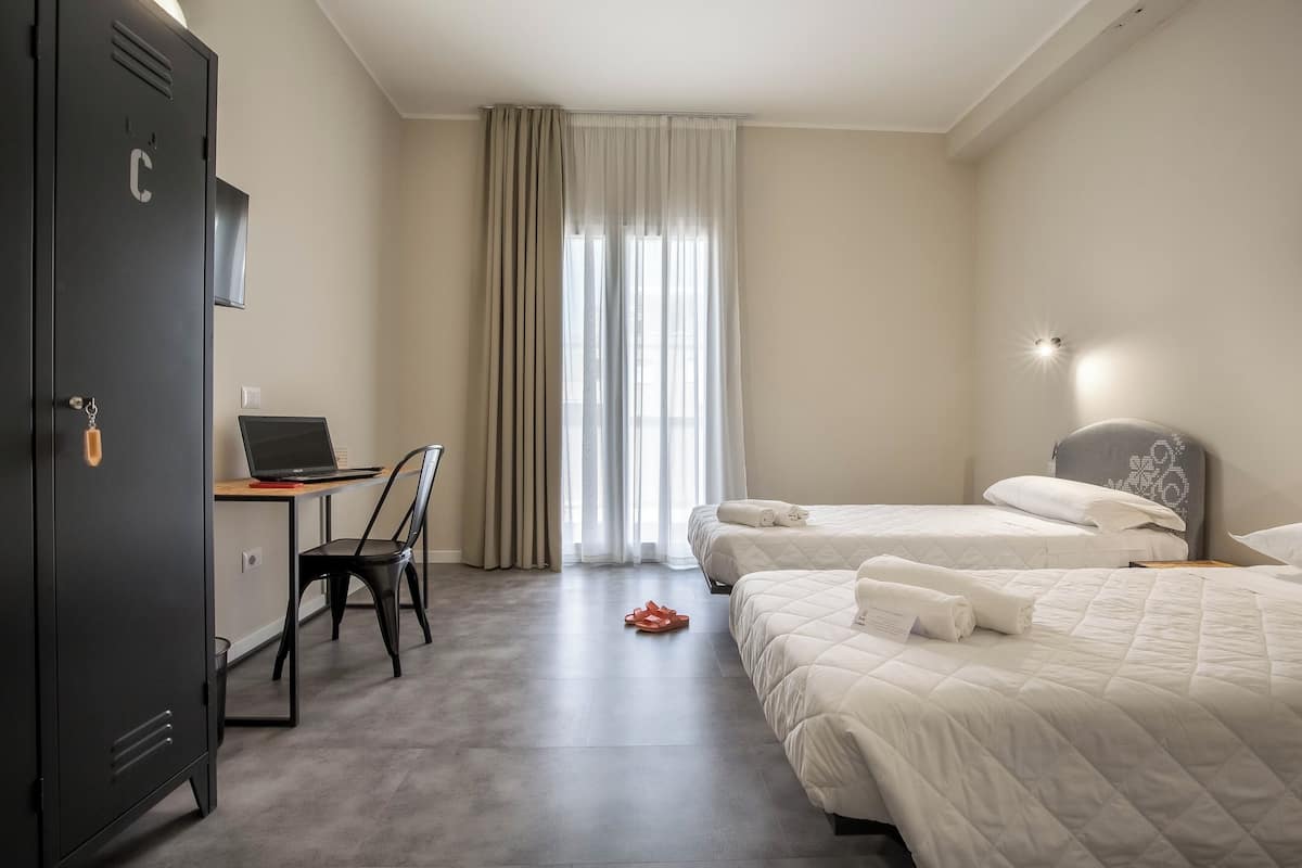 A hotel room with a desk, two beds, a wardrobe, and a large window with curtains.