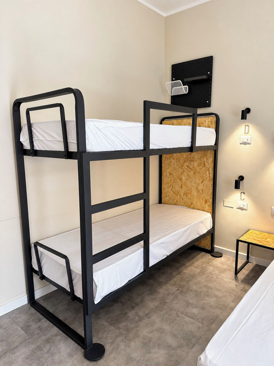 Una cama litera con cabecera de madera, una mesita de noche pequeña y pulsadores de luz en la pared.