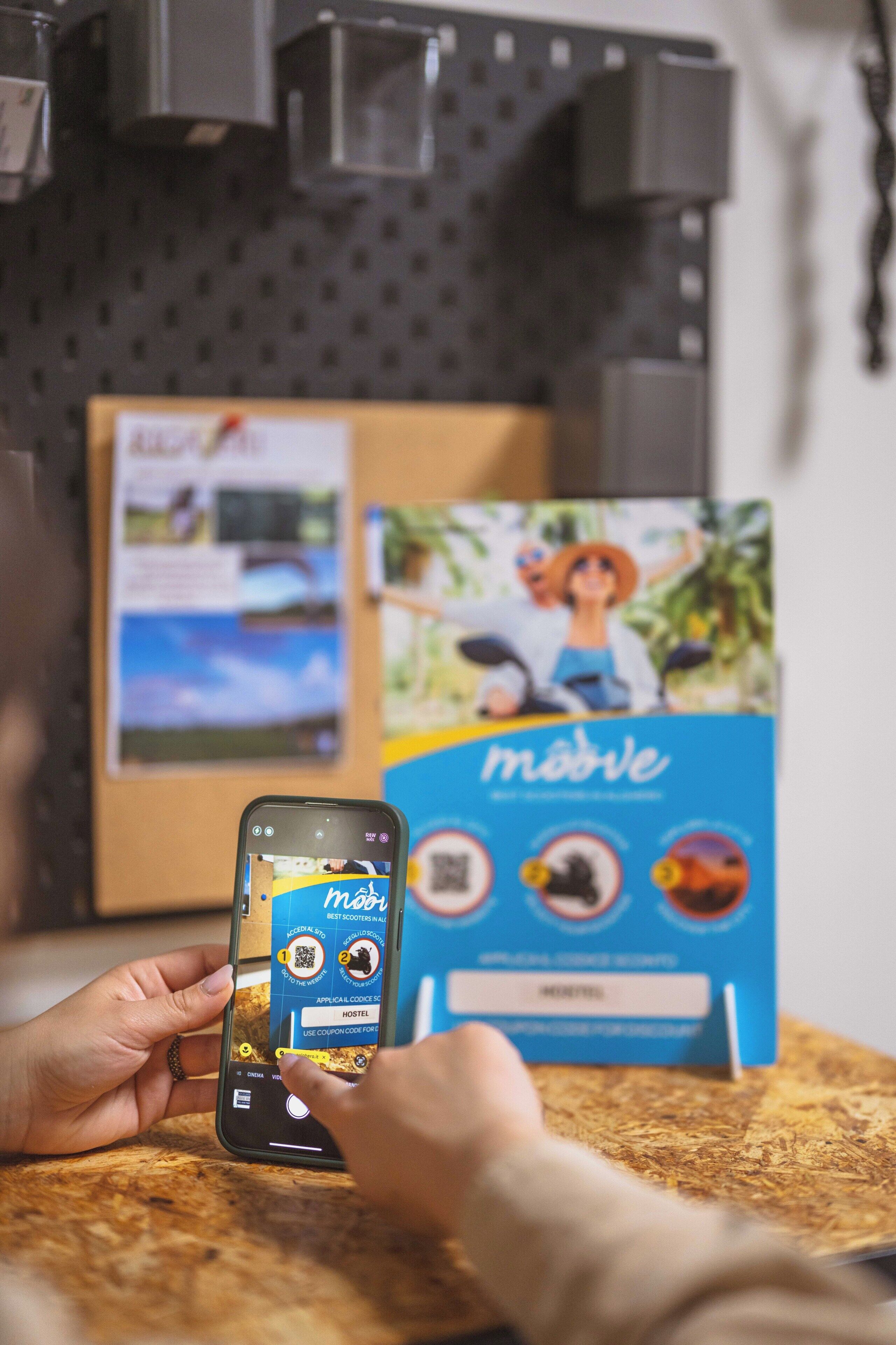 A person using a smartphone with a travel app interface, a cardboard box with a travel-related image, and a pegboard wall in the background.