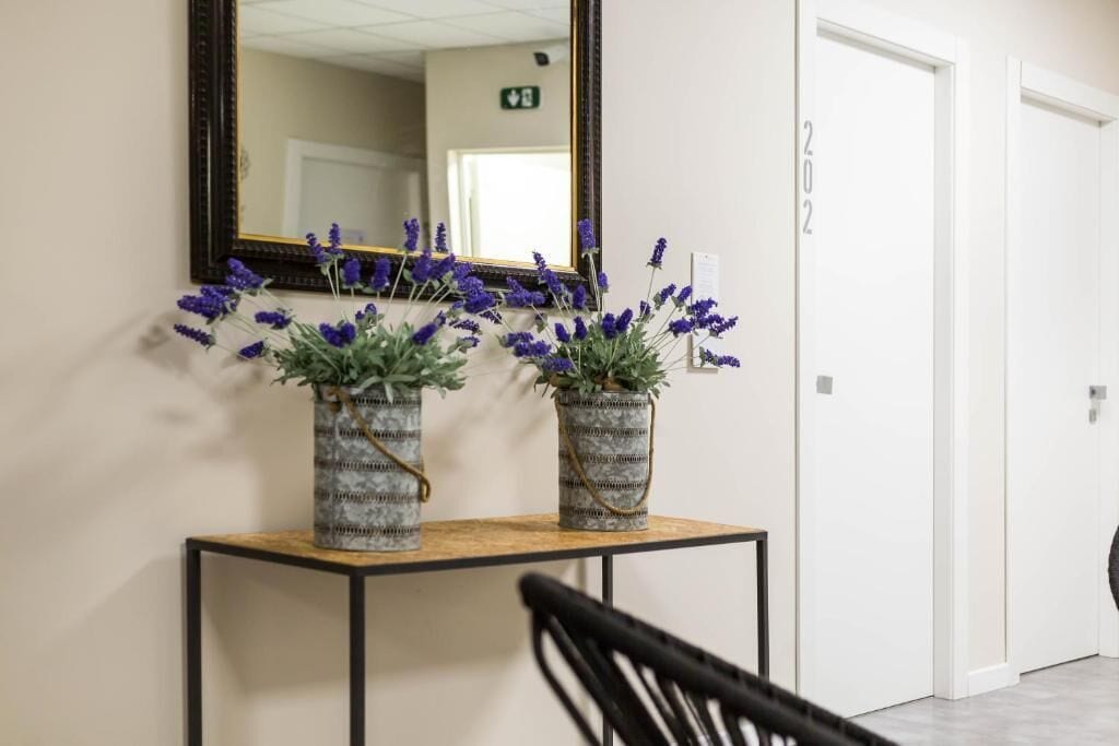 Un pasillo con una consola de madera, dos macetas de lavanda, un espejo y una puerta blanca con el número 202.