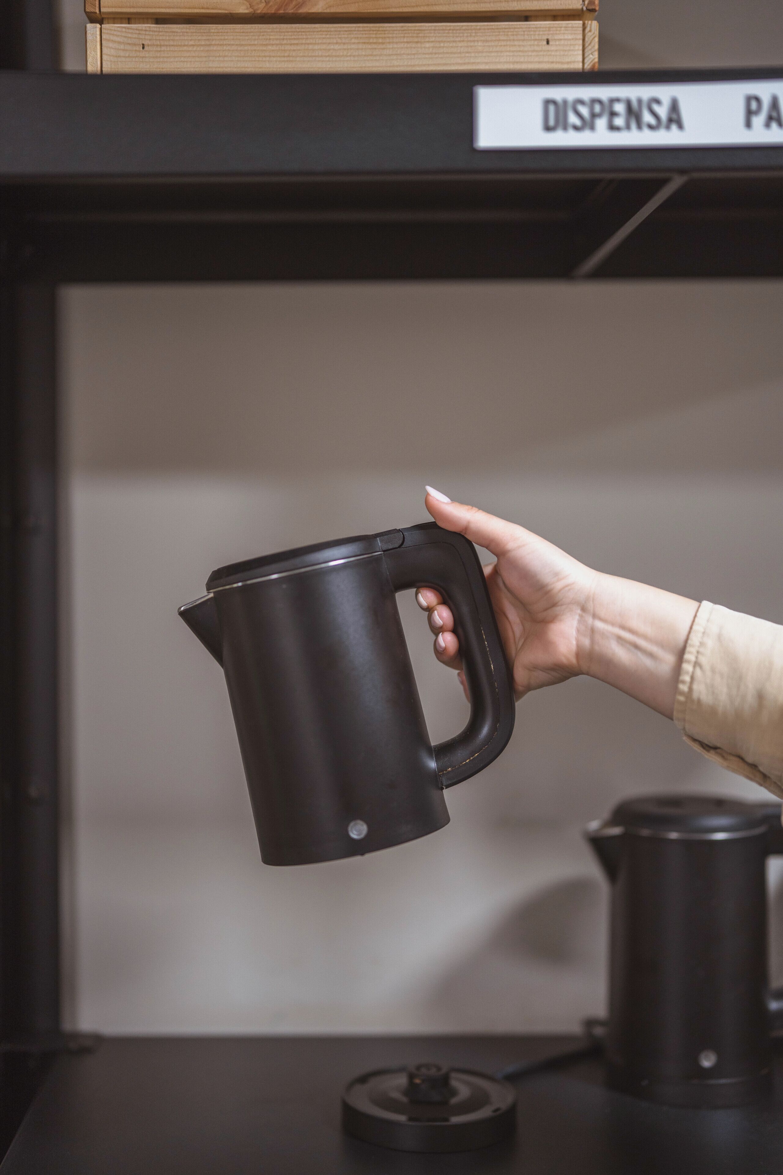 Eine Hand hält eine schwarze elektrische Wasserwaage auf einem Tisch mit einer Kaffeemaschine und einem Schild, das einen 'DISPENSA'-Bereich ausweist.