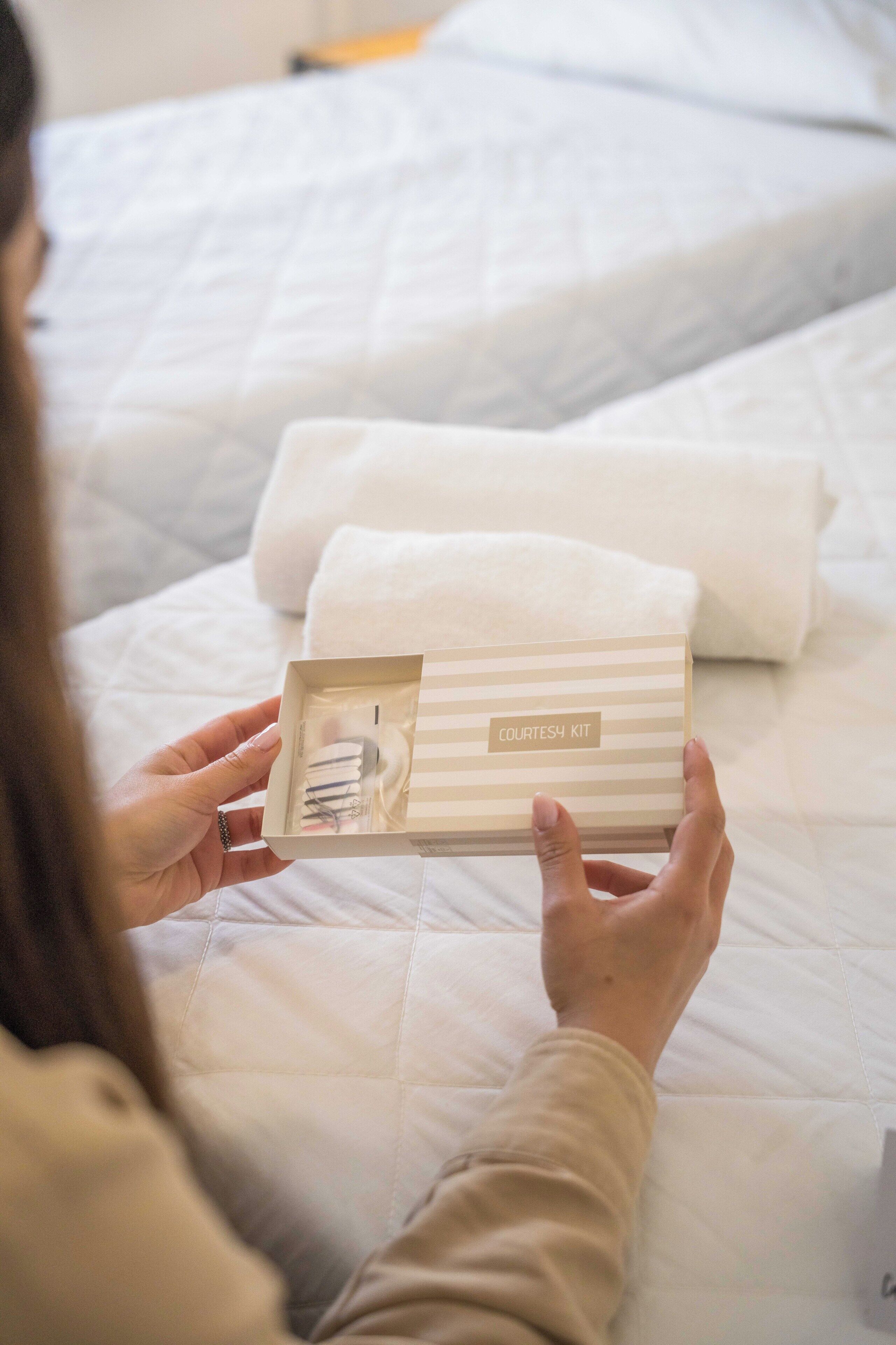 A person holding a box labeled 'COURTESY KIT' on a bed with white bedding.