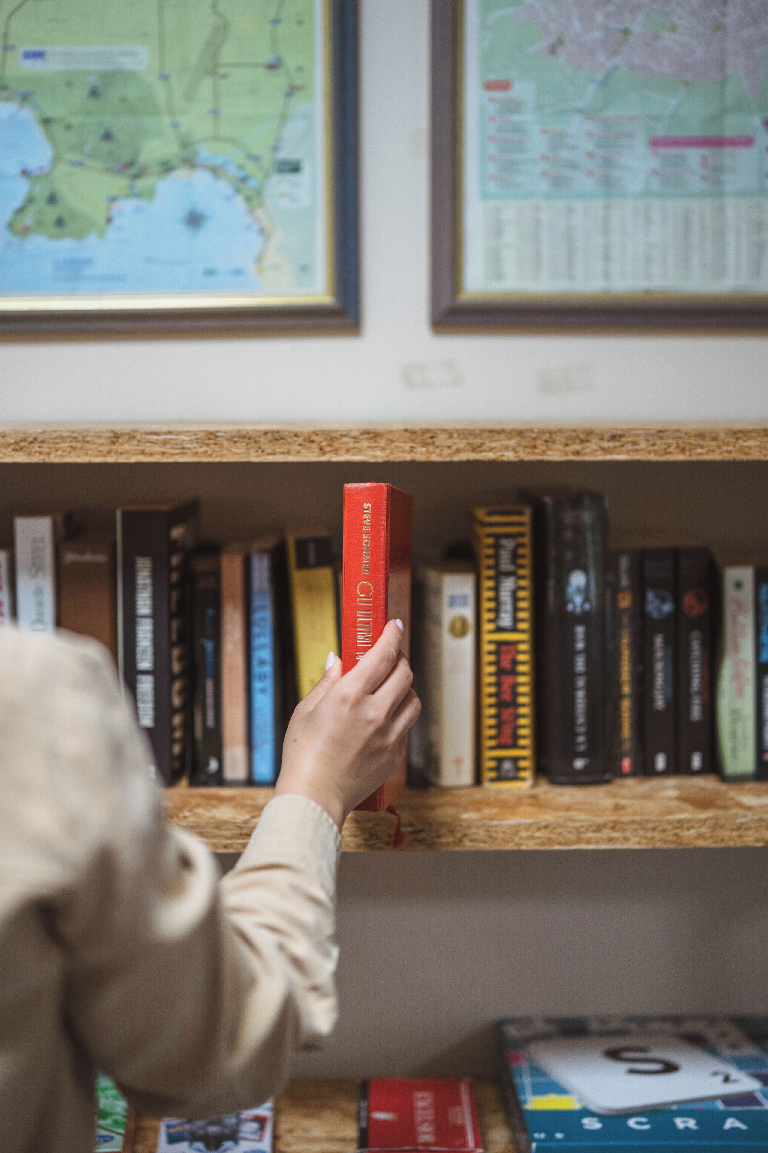 A hand holding a red book on a bookshelf with various other books and a framed map on the wall.