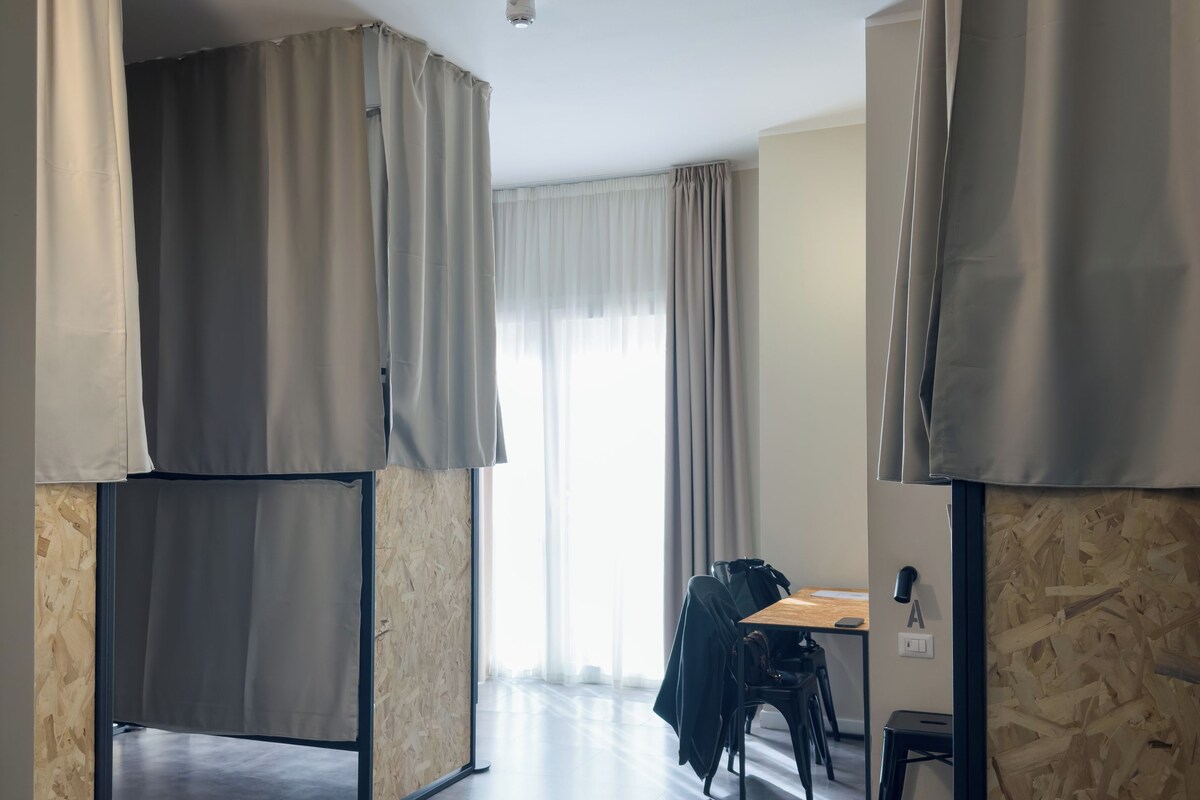 A modern, minimalist room with a wooden floor, a desk with a chair, and a large window with curtains.