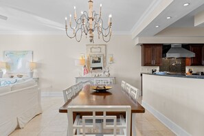 A spacious dining area with a wooden table and white chairs, a chandelier, and a kitchen area with dark wooden cabinets and a modern stove. - Amazing Condo on the Beach on 30A! (Santa Rosa Beach)