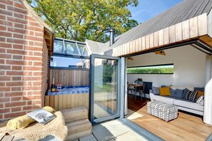Un espace de vie extérieur moderne, doté d’une porte vitrée donnant accès à un patio, d’une terrasse en bois et d’un coin salon confortable agrémenté de coussins et d’un pouf à motifs.