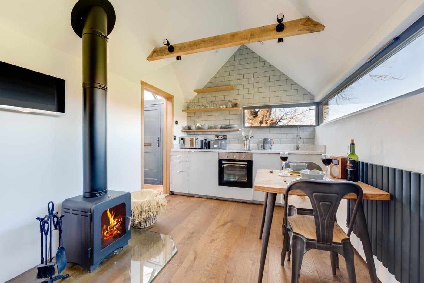 A modern kitchen with a wood-burning stove, a dining area with a table and chairs, and a view of the outdoors through a window.