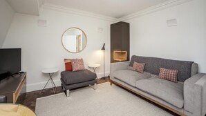 Un salon moderne avec un canapé gris, un petit pouf, un miroir rond, un téléviseur posé sur un meuble en bois et une lampe sur pied.