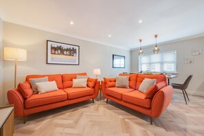 Ein modernes Wohnzimmer mit zwei orangefarbenen Sofas, einem Essbereich mit Tisch und Stühlen sowie einer Küche im Hintergrund.