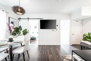 A modern living room with a glass dining table, white chairs, a black sofa, and a large flat-screen TV mounted on the wall. - Cozy Home w/ parking, Walk to Clifton Hills, 10mins to The Falls, Fire Pit &more (Niagara Falls)