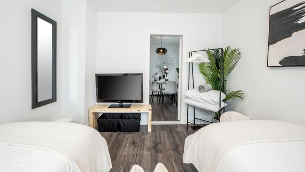 Una moderna camera d'albergo con due letti, una TV, una scrivania con un computer, un'area pranzo e una pianta di grandi dimensioni.