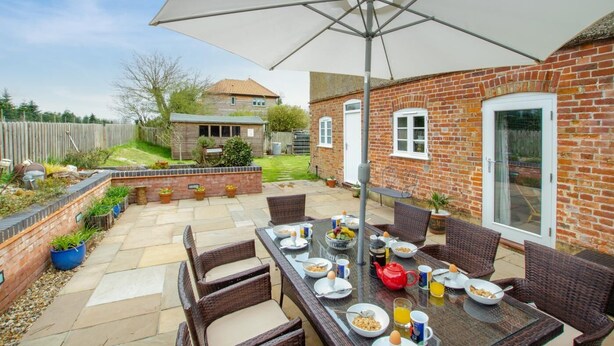 Une terrasse extérieure avec une table à manger dressée pour le petit-déjeuner, entourée de chaises en osier et d’un grand parasol.