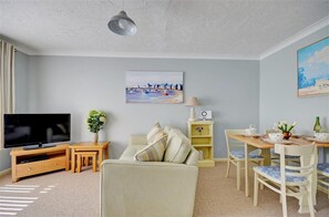 A living room with a TV, sofa, dining table, chairs, and a painting on the wall. - 2 Bed in Wells-next-the-Sea (oc-1655) (Wells-next-the-Sea)